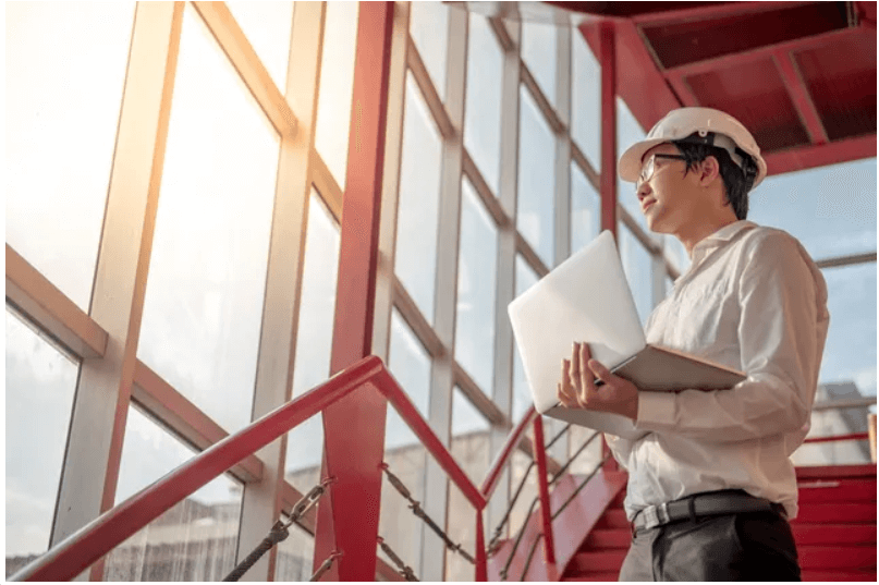 Engineer with laptop stands by large windows.